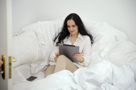beautiful black-haired woman sits on white sofa near a duvet in hands with tablet corner of room works plays communicates nearby mobile phone social networks Internet Call friend pleasant emotionsの写真素材