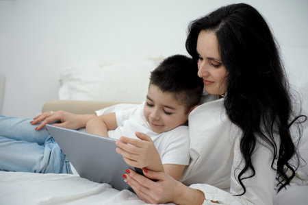 mom and son are lying comfortably on the bed relaxing playing a game on a tablet calling home have gone to another country white clothes black hair love caring family relationships motherhoodの写真素材