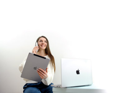 new technologies Apple MacBook 14 pro phone iPhone 13 pro Max and iPad on a white background the girl uses all the latest technologies talking on the phone smiling holding a tablet looking at a laptopのeditorial素材