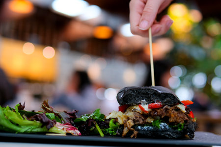 hand take out a skewer from a black burger bun with ink Big cheeseburger with lots of cheese. Stock photo side view of a cheeseburger on a black brick wall background.の写真素材