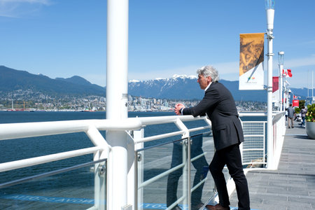 handsome gray-haired man in suit stands by water looks at mountains and nature tranquility peace vacation in Vancouver Ocean lean on handrail canada place sail on ship office worker director managerのeditorial素材