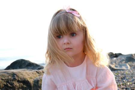 a small child a girl in a pink delicate dress sits, on a stone and looks into the distance blonde curly hair childhood loneliness sea sunset there is a place for textの写真素材