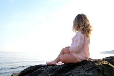 a small child a girl in a pink delicate, dress sits on a stone and looks into the distance blonde curly hair childhood loneliness sea sunset there is a place for textの写真素材