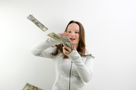Arrogant wealthy girl scattering dollars with proud haughty expression, boasting rich life, throwing around cash, squandering wasting money carelessly. indoor studio shot isolated on blue backgroundの写真素材