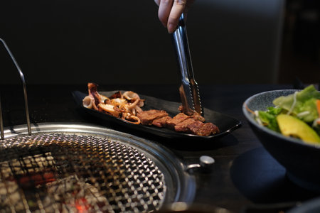Red king prawns sizzle on a hot grill beside thick beef steaks. A cook uses metal tongs to flip the meat on the grate. Juicy seafood and beef grilling over open fire a delicious mix of surf and turf.の写真素材