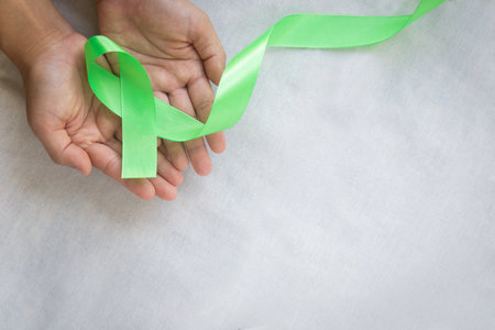 Hands holding emerald green or jade green ribbon on white fabric background with copy space, symbol for Liver Cancer awareness, World Cancer Day. Healthcare or hospital and insurance concept.の写真素材