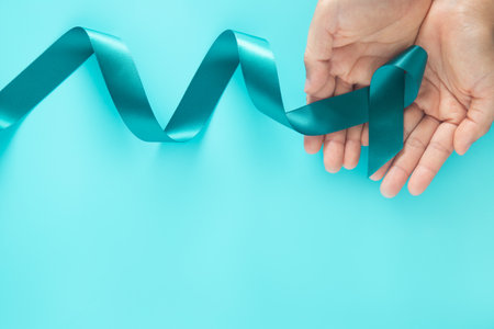 Hands holding teal color ribbon on teal background with copy space. Ovarian Cancer Awareness, Gynecological, Uterine Cancer, Vulvar Cancer, Panic Disorder, Post Traumatic Stress Disorder (PTSD).の写真素材