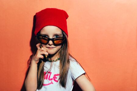 Little hipster girl stands against the wall and looks out from under the glasses and speaks on the phoneの写真素材