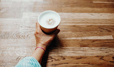 Female hand holding a paper cup with coffee on a wooden table, top viewの写真素材