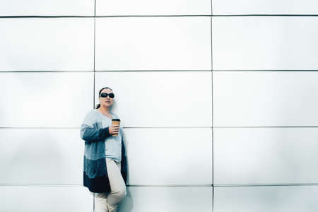 A woman in sunglasses stands with a glass of coffee standing against the wall