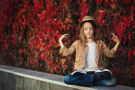 A child sits in a lotus pose against the background of autumn leavesの写真素材