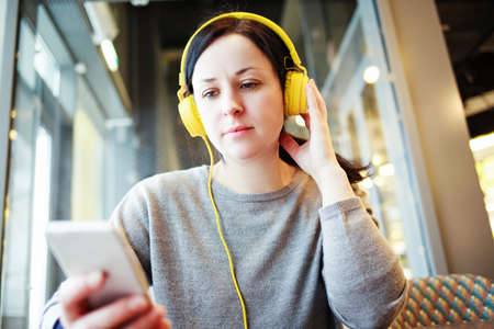 Woman in yellow headphones working on a smartphone in a cafeの写真素材