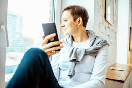 Woman sitting on windowsill with smartphone, working remotely during quarantine