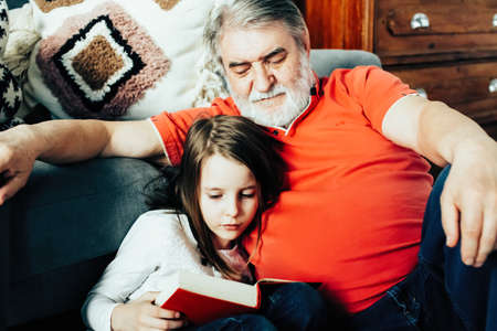 Granddaughter reads a book to her grandfather at homeの写真素材