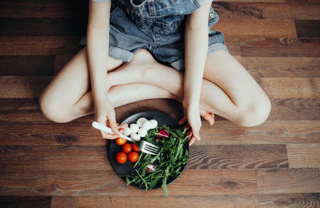 Green healthy breakfast i salad, tomato and mozzarella for child. Girl eats sitting on the floor, top viewの写真素材