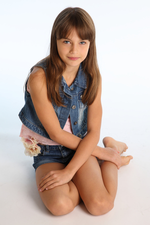 Beautiful girl in a denim shorts is resting on the floor barefoot. Elegant attractive child with a slender body and bare long legs. The young schoolgirl is 9 years old.