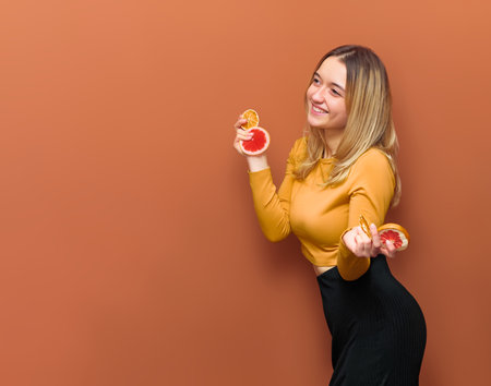 Young attractive girl holds grapefruit and orange slices in her hands, smiling broadly looks into copy space, in yellow blouse and black bottom on vanilla background.の写真素材