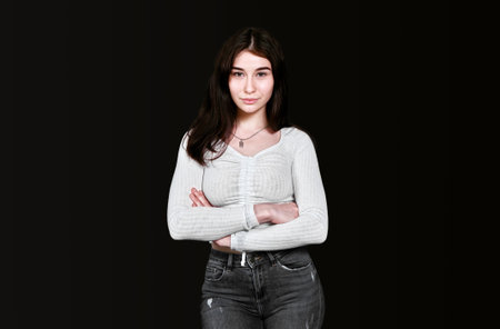 Slender young brown-eyed brunette in white blouse and black denim trousers crossed her arms over her chest, standing and looking at camera with smile on black background. Banner, place for text.の写真素材