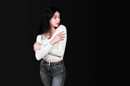 Slender young brunette in white blouse and black denim trousers crossed her arms over her chest, standing and looking at her shoulder against black background. Banner, place for text.の写真素材