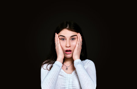Portrait of surprised young girl with long black hair looking at camera with open mouth on dark background with space for text. studio shot.の写真素材