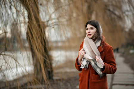 Young attractive brunette girl, with scarf around her neck, in orange coat, holding scarf with her hands, looking away, standing in autumn park against backdrop of blurry trees and river, copy space.の写真素材