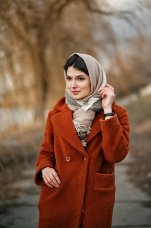 Young brunette girl in orange coat and woolen scarf straightens her headscarf with her hand, looking away, standing against backdrop of autumn park in blur.の写真素材