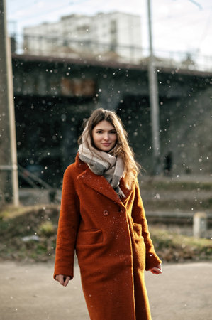 Young girl in orange coat with scarf around her neck, posing with her hands down, standing under falling snow, against background of bridge in blur, looking at camera, with copy space.の写真素材