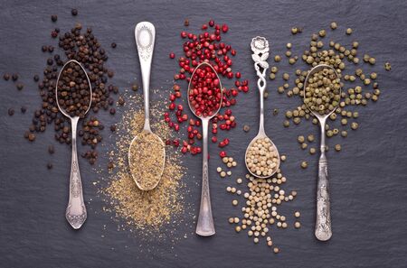 Various kinds of peppercorns in silver spoons on black stone background
