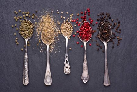 Various kinds of peppercorns in silver spoons on black stone backgroundの写真素材