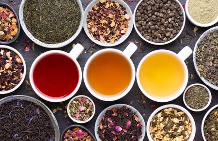 Assortment of tea cups, dried leaves, fruit and herbs in bowls on dark, stone background, top viewの写真素材