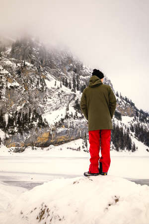 Man looking to the horizon in front of a frozen lake in a snowy mountain landscape on a cloudy day in Switzerlandの写真素材