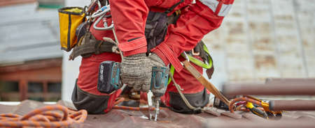 Roof repair. Installation of a metal roof. A man in a red suit works on a red roof. Work at a height. rope access.の写真素材