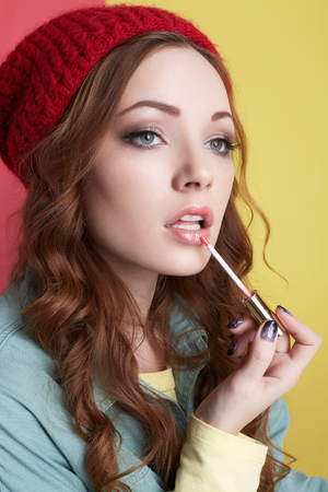 fashionable hipster girl in hat, painting lips with lipstick. Beautiful young woman applying makeup. winter fashion. Beauty Model Girl with curly hair and red capの写真素材