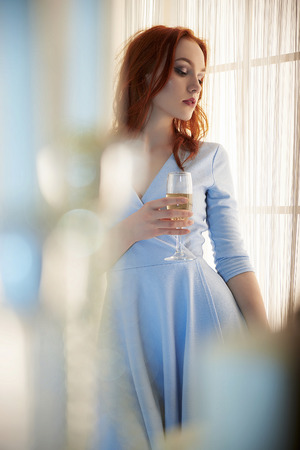 beautiful young woman drinking champagne, stay near the window. winter home lifestyle. Happy New Year party. beauty red haired girlの写真素材