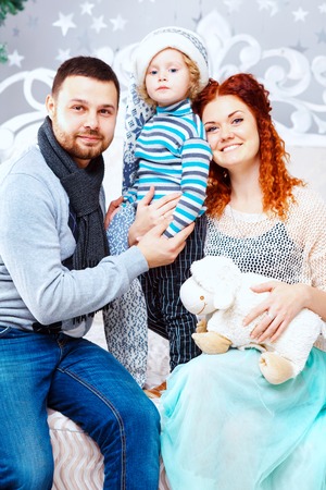 Christmas family of three persons mother father and son two year old boy and fir tree with gift boxes over white bedroom backgroundの写真素材