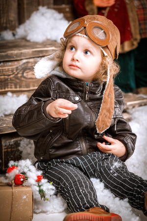 Little two year old boy dressed in braun leather jacket, pants and boots with pilot hat on posing plays with snow in christmas wooden decorationsの写真素材