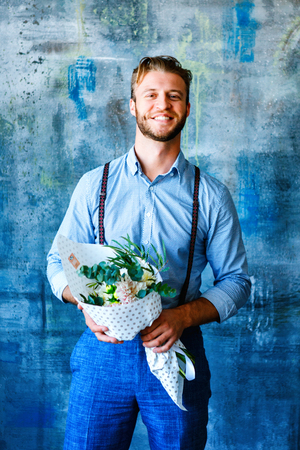 Male positive smiling florist man holding bouquet with flower orange roses on blue background, wearing blue pants and shirtの写真素材