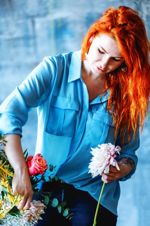 Charming cheerful female shop assistant making bouquet with purple crocus and white roses. Flower shop and floristic design master class concept on painted blue wall loft styleの写真素材