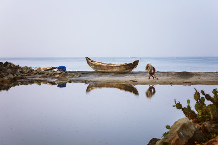 Varkala, India - February 09, 2016: Fisherman carrying net ropes walking on the sand, Wooden fishing boat anchored on the beach, reflection in backwaters of Odayam beach near Varkala, Kerala, Indiaのeditorial素材