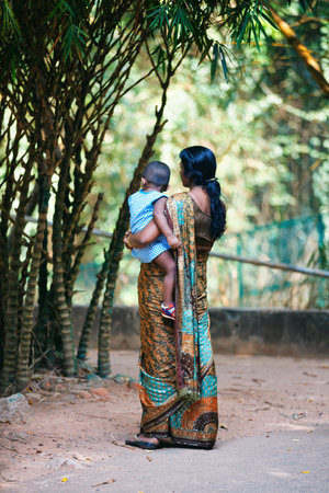 Trivandrum, India - February 17, 2016: Indian mother wearing female sari holds little child, view from the back, on green park backgroundのeditorial素材