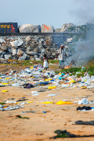 Kollam, India - February 14, 2016: A child girl plays in piles of trash while her mother burns it on the beach of Kollam, Keralaのeditorial素材
