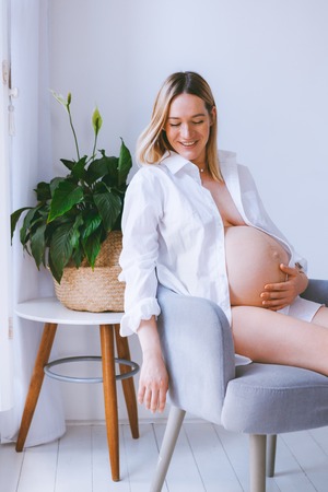 Pregnant mothercare lifestyle concept, Bob hairstyle blond woman health care. white light interior, mother sitting on the grey armchair in the room with blinders and wooden floor. green plant in the cornerの写真素材