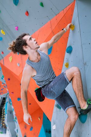 Urban concept of man with curly long hair, tied in a ponytail, is training at the city artificial red and blue climbing wall using talcum powder, wearing a bag for climbing without insurance equipmentの写真素材