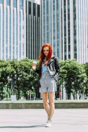Happy young trendy red hair woman wearing black leather jacket and shorts, drinking take away coffee and walking in an urban city. City walk lifestyle concept.の写真素材
