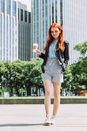 Happy young trendy red hair woman wearing black leather jacket and shorts, drinking take away coffee and walking in an urban city. City walk lifestyle concept.の写真素材