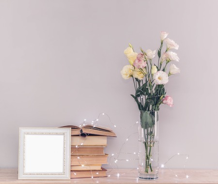 Eustoma flowers bouquet in a vase, stack of old vintage books, white photo frame and garland lights on a grey background. Reading and relax conceptの写真素材