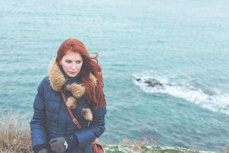 Girl in a warm jacket with fur stands on the frozen bank of the winter sea