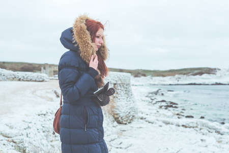Girl in a warm jacket with fur stands on the frozen bank of the winter sea