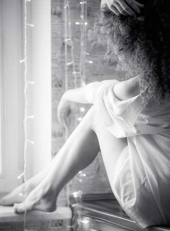 Blurred portrait of a curly girl with bare feet sitting in a men's white shirt by the window. No face, BW photo.の写真素材
