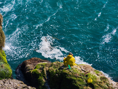 A man in a yellow raincoat sits on a cliff and fishes in the sea near a waterfall in Turkey. Turquoise sea. View from above.の写真素材